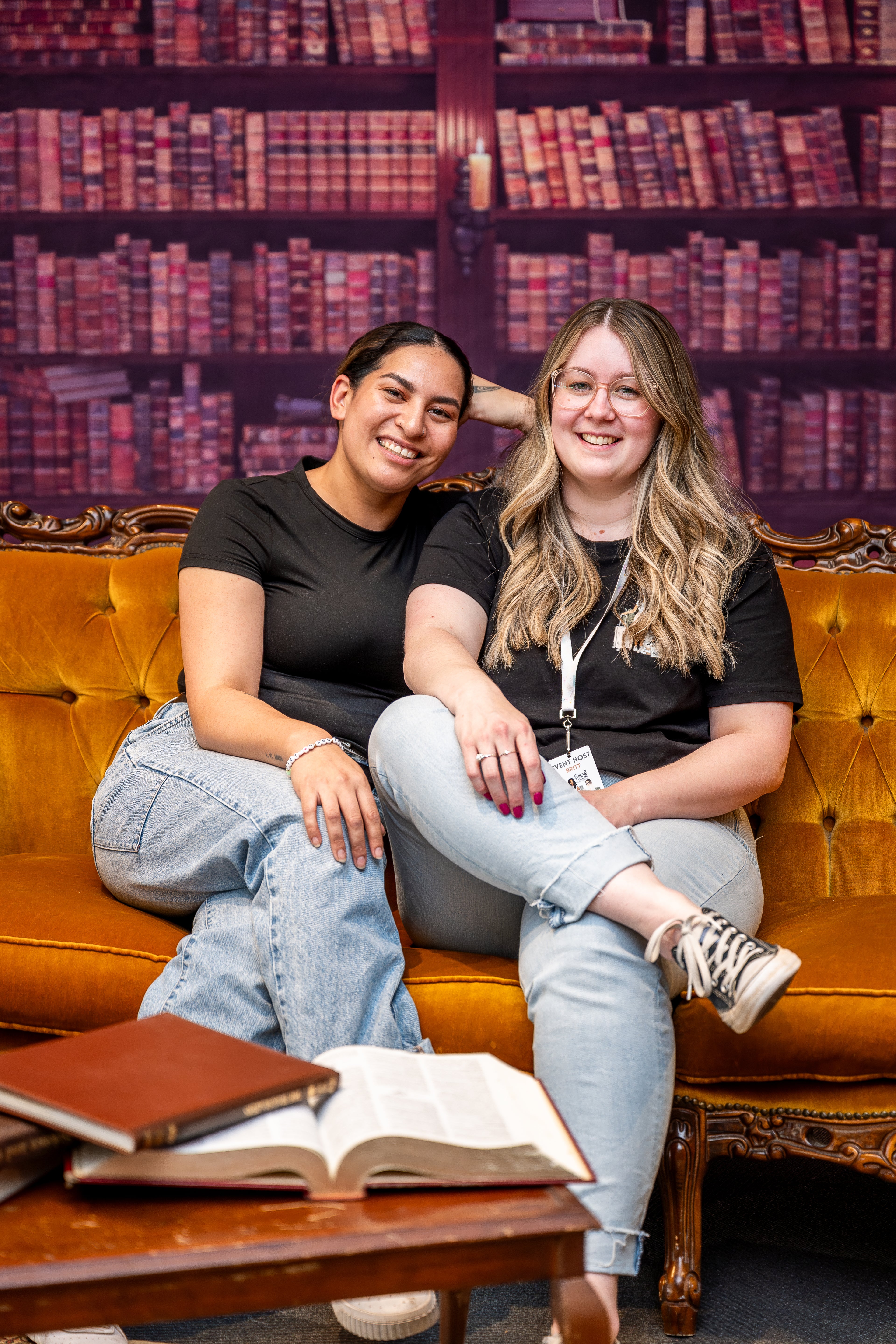 Two women sitting on a couch in a library with bookshelves in the background.